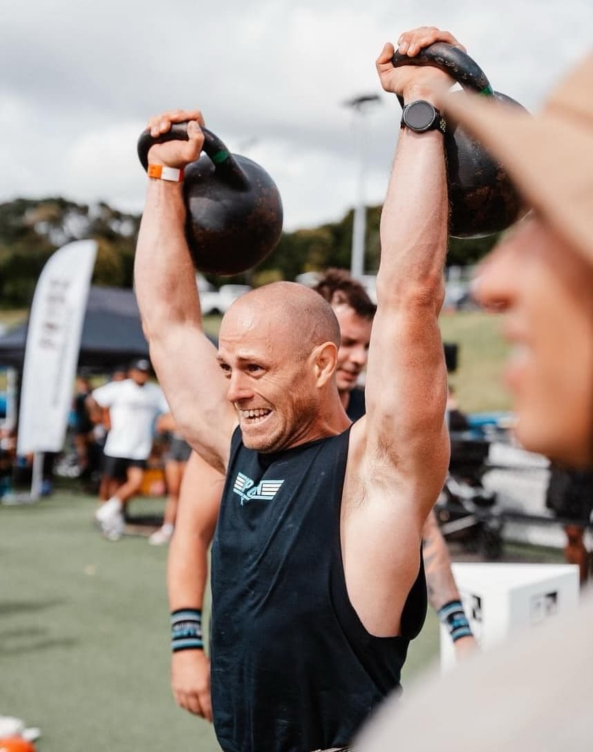 A photo of Coach Ryan doing an overhead press with kettlebells in a CrossFit competition